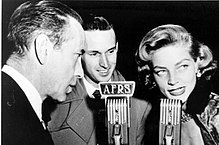 Lauren Bacall in black and white with Humphrey Bogart speaking into microphones