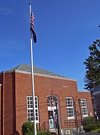 A small brick building with hipped roof and a flagpole in front seen from its left