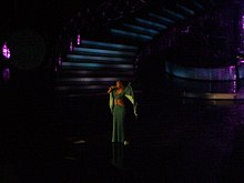 A woman is performing a song on stage. She is wearing a greenish dress.