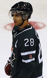 Hockey player in black uniform, with the number 28 on his shoulder. He stands on the ice, facing to the left of the camera.