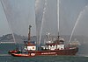 A red fireboat at rest on calm water sends five jets of water into the air.