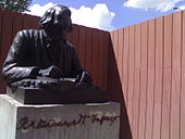 Outdoors bronze statue (depicting Tagore at a desk and writing in a book) on a stone plinth with inscribed cursive name.