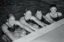A black-and-white photograph of four girls in a swimming pool all looking at the viewer and smiling while holding onto the side of the pool