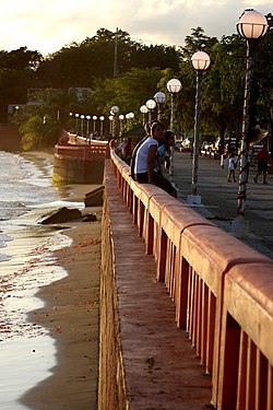 Пляж слева, моющийся до променада, люди сидят на стене спиной к океану, длинная очередь уличных фонарей справа