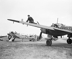 A photograph of a single–engined and a larger, twin engined aircraft on the ground. Two men are working on the wing of the larger aircraft. The engine–covers have been removed from the smaller aircraft and four men are working on its engine