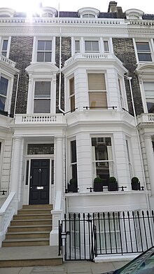 Élégante maison de ville mitoyenne, sur trois étages, aux murs et fenêtres blancs. La partie supérieure de la maison est en briques brunes. Un petit escalier mène à la porte d'entrée.