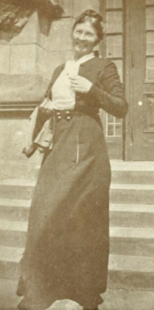 A white woman wearing a dark suit with a short jacket and long skirt, smiling, outdoors, in front of a building's steps