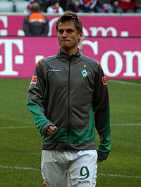A photograph of a blonde man wearing a grey and green training jacket and white shorts, the man is seen on a football pitch in the process of warming up.