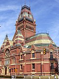 An ornately decorated brick and stone building with a multicolored slate roof. It is two stories, but has a very steep roof that almost doubles the building's height, and there is a tower rising even higher. Turrets flank the main doorway, above which is a stained glass window.