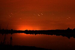 A red sky reflected in a lake at night, with short time-lapse trails of brighter stars