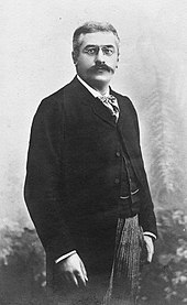 Photo en noir et blanc longueur trois-quarts d'un homme se tenant debout, avec les cheveux grisonnants, des sourcils et une moustache foncés, portant des lorgnons, un foulard, une chemise blanche, un veston et une redingote noirs, un pantalon clair.