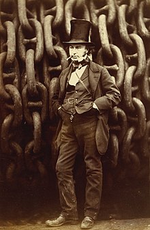 A 19th century man wearing a jacket, trousers and waistcoat, with his hands in his pockets and a cigar in mouth, wearing a tall stovepipe top hat, standing in front of giant iron chains on a drum.