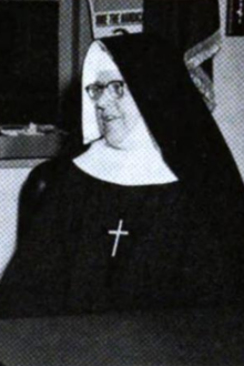 A white woman wearing glasses and a traditional nun's habit, with wimple headwear and a cross on her chest