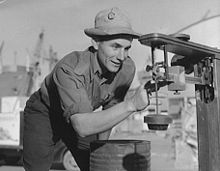 A smiling man in a short-sleeved shirt, dark pants, and a light-colored bucket hat examining a piece of mechanical equipment