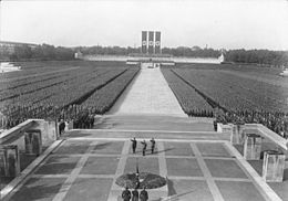 A distant photograph of a Nazi rally, with large crowds on either side of a long empty stretch with Nazi flags at the end.
