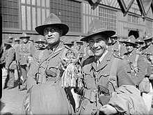 Two Maori officers wearing military uniforms smile at the camera, surrounded by other soldiers in front of a building