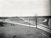 "A black and white photograph of a divided roadway being crossed by another roadway. The horizon cuts the photo in half. The setting is mostly rural, with the roadways slicing through that. The divided road extends from the lower right corner to the centre of the horizon, while the second roadway crosses horizontally halfway towards the foreground. Connecting the two separated roadways are a series of ramps. Although only half visible from the angle of the photo, the ramps form the shape of a four-leaf clover surrounded by a diamond."