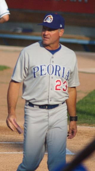 A man wearing a gray baseball uniform with "PEORIA" across the chest and a blue baseball cap walking on a grass field