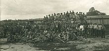 Photo noir et blanc d'un grand nombre d'hommes sur plusieurs rangs devant des baraquements en bois.