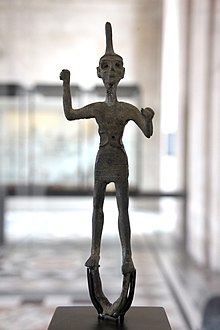 A bronze statue of a standing male figure, his right hand raised and his left hand extended, wearing a crested helmet