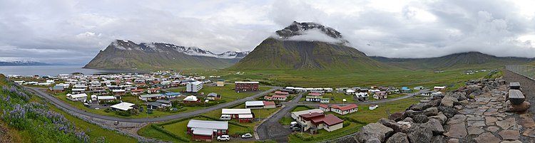 A small village of white-roofed buildings sits in front of steep green mountains, with their tops obscured by clouds. An ocean bay is visible to the left of the image, and purple flowers dot the hills in the foreground.