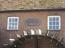 Brick building with tiled roof and two square windows, below which is a water wheel. Between the windows there is a wooden plaque, which reads "Cobham Mill Restored 1993 by Cobham Mill Preservation Trust and the National Rivers Authority".