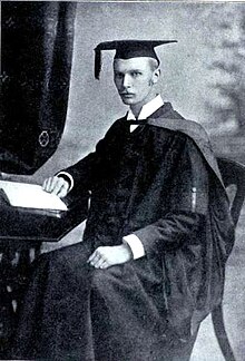 Young man wearing a cap and gown, seated in front of a small table with a book on it