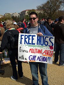 Smiling man in sunglasses at an outdoor rally, displaying a hand-lettered sign which reads, "Free Hugs from a Militant Atheist with a Gay Agenda"