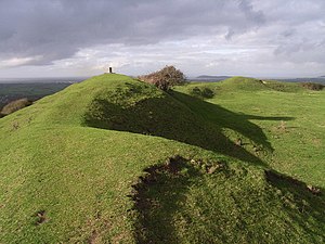 A photograph showing the hill fort called "Brent Knoll Camp" under a partly cloudy sky. The hill fort is on the top of the hill, and banks can be seen circumscribing the plateau.