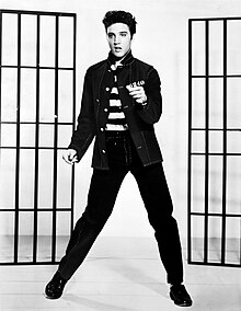 A young man dancing, swiveling his hips. He has dark hair, short and slicked up a bit. He wears an unbuttoned band-collared jacket over a shirt with bold black-and-white horizontal stripes. Behind him, on either side, are a pair of barred frames, like prison doors.