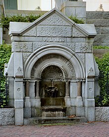 A small water fountain with a gothic structure along a brick sidewalk, flanked by wrought iron fences