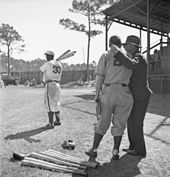 Two white men in baseball uniform with back to camera watch a black baseball player take batting practice