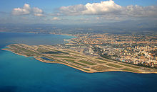 Farbfotografie in der Vogelperspektive von einem Flughafen mit Landebahnen, die links an drei Seiten vom Meer umgeben sind. Rechts ist eine große Stadt und im Hintergrund ist eine Berglandschaft.