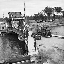 Photo en noir et blanc d'un camion passant sur un pont métallique gardé par plusieurs soldats et une Jeep.