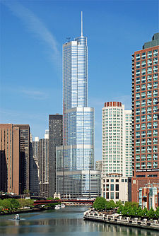 A tall silver skyscraper sits at a jog in the river beyond a bridge. The river and other along its banks buildings are in the foreground.