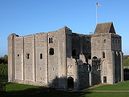A square building of grey stone with narrow vertical slits on the first floor, and wider windows on the second. The top of the castle looks decayed and there is no roof, except over a tower attached to the keep.