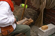 Foto einer von schräg hinten aufgenommenen Person beim Hecheln von Flachsfasern. Im Bildausschnitt sind von der Person nur Oberkörper und Oberschenkel zu sehen. Sie sitzt auf einer Bank. Vor ihr liegt ein Brett, aus dem auf einer Fläche von etwa 10 mal 20 Zentimeter Nagelspitzen ca. 10 cm hoch hervorragen. Diese bilden eine Art Kamm. Durch den Kamm zieht die Person mit beiden Händen in horizontaler Richtung ein Bündel Flachsfasern auf sich zu.