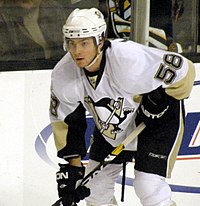 A hockey player in white uniform. He crouches, holding his stick in a ready position.