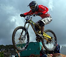 Ein Mountainbikefahrer springt mit einem Downhill-Mountainbike. Er trägt ein rotes, flatterndes Trikot.