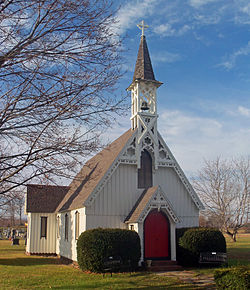 Белая церковь с вертикальной обшивкой, декоративной деревянной обшивкой по линии крыши круто остроконечной крышей и красной дверью впереди, увенчанной открытой колокольней.