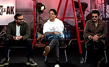 Three men are seated in a row in from of various film set equipment and a sign reading "AK vs AK".