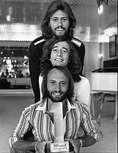 Three long-haired men, two with beards, standing together holding a trophy shaped like the number one