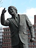 A bronze statue of a man in a suit, pointing with his right hand.