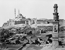 Многокуполна джамия доминира в оградената цитадела, с разрушени гробници и самотно минаре отпред.