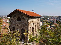 Southwest view of a narrow medieval church topping a cliff, with a cityscape in the background