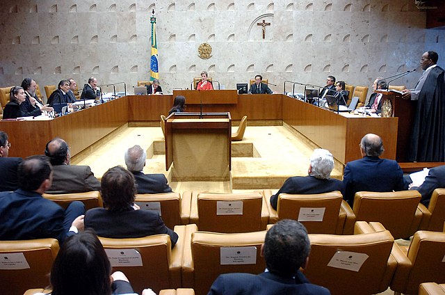 Em primeiro plano, as cadeiras de espectadores do julgamento, todos trajados formalmente. Ao fundo e centralizada, a presidente do STF Ellen Gracie; à direita dela, o PGR e a sua esquerda um assessor judiciário. Atrás dela, a bandeira do Brasil, o emblema nacional e um crucifixo cristão. Os outros ministros se dispõem lateralmente. A direita, o ministro Barbosa está de pé, em frente de um púlpito com microfone, lendo.