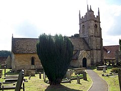 Bathstone building with prominent four stage tower at the right hand end. Partly obscured by a yew tree.