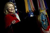 Хилари Клинтон носи црвени мантил и стоји за говорницом на којој је печат Министарства одбране САД; она је насмејана и прича у црни микрофон, окренута надесно и с десном руком наслоњеном на говорницу