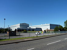 A photograph of a modern factory with two large industrial buildings standing side by side and a loading area at the front. The name of the company is written in red lettering at the front of each building. A column of steam rises in the background