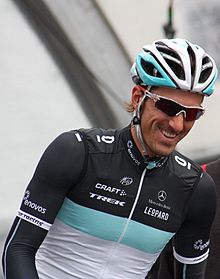 A road racing cyclist wearing a jersey with black sleeves, a white torso, and a blue stripe separating them. He is smiling, and also wears sunglasses with red lenses and a blue and white helmet.